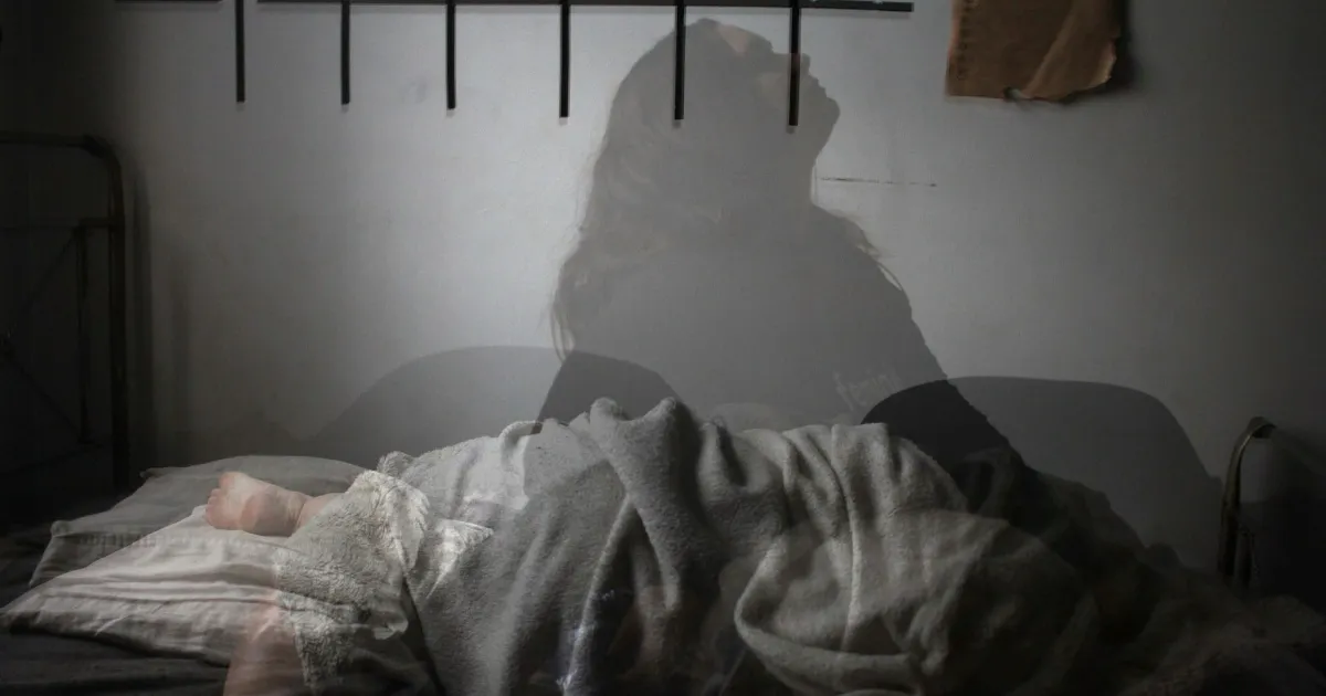 A man and woman wrapped in sheets in low light, his gaze drifting past her