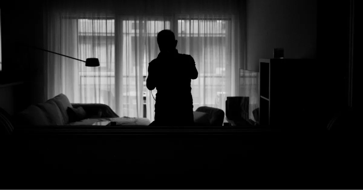 A man standing alone at a bedroom window at night, the empty bed behind him