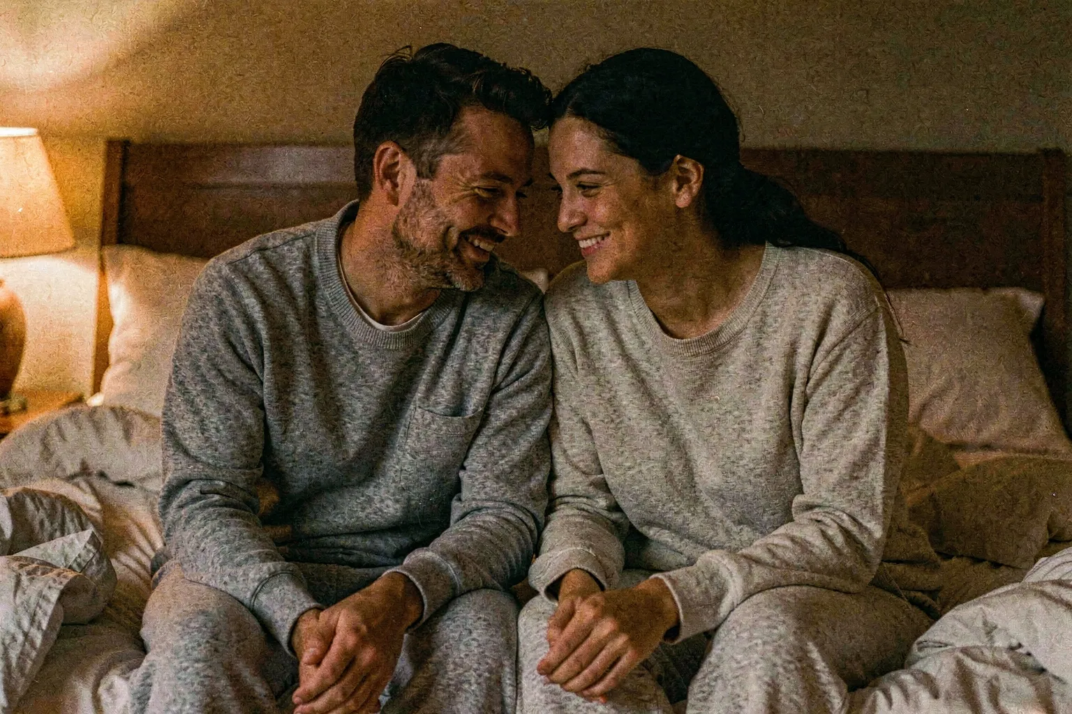A couple sitting on the edge of a bed, fully clothed, leaning close in warm lamp glow, one softly laughing and the other smiling warmly, the sheets rumpled around them