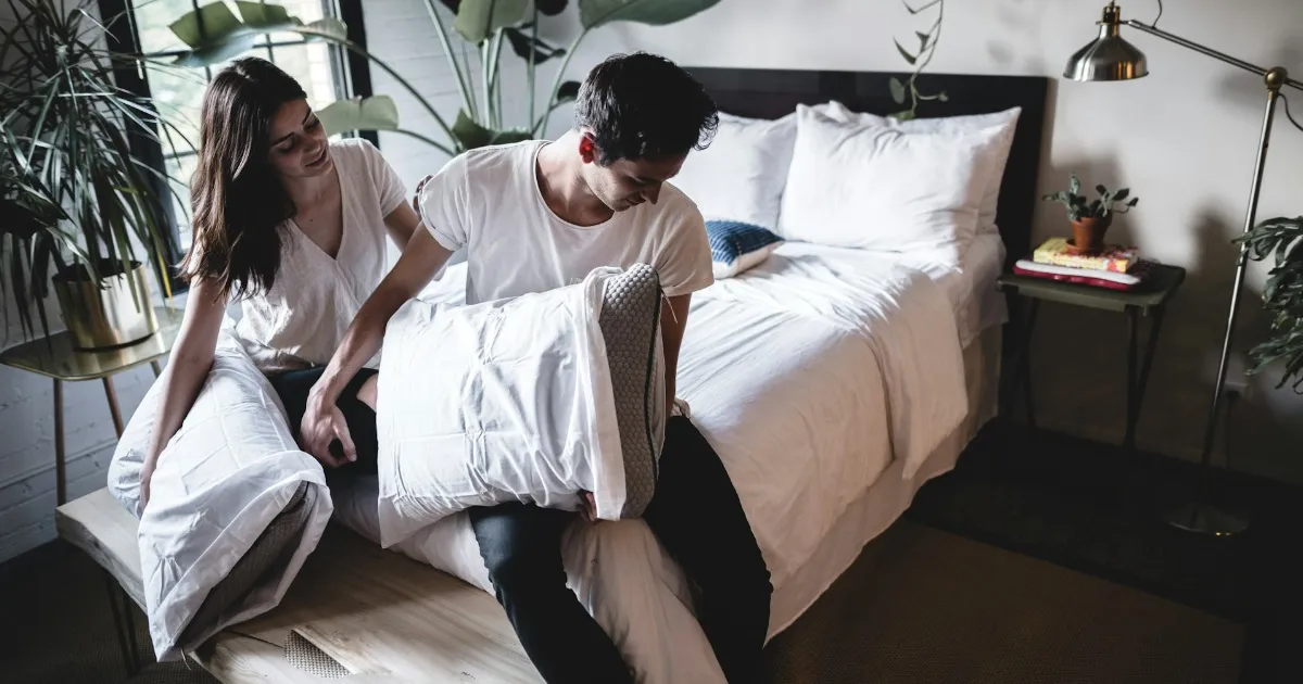 A couple mid-laugh in a rumpled bed, her hand on his chest pushing back as he leans in, warm lamplight on the sheets