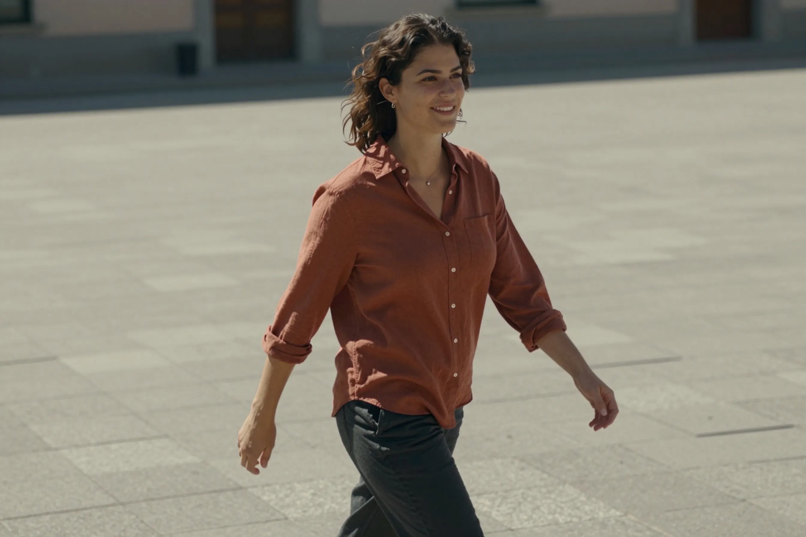A woman in her late 20s walking with confident posture across a sunlit stone plaza