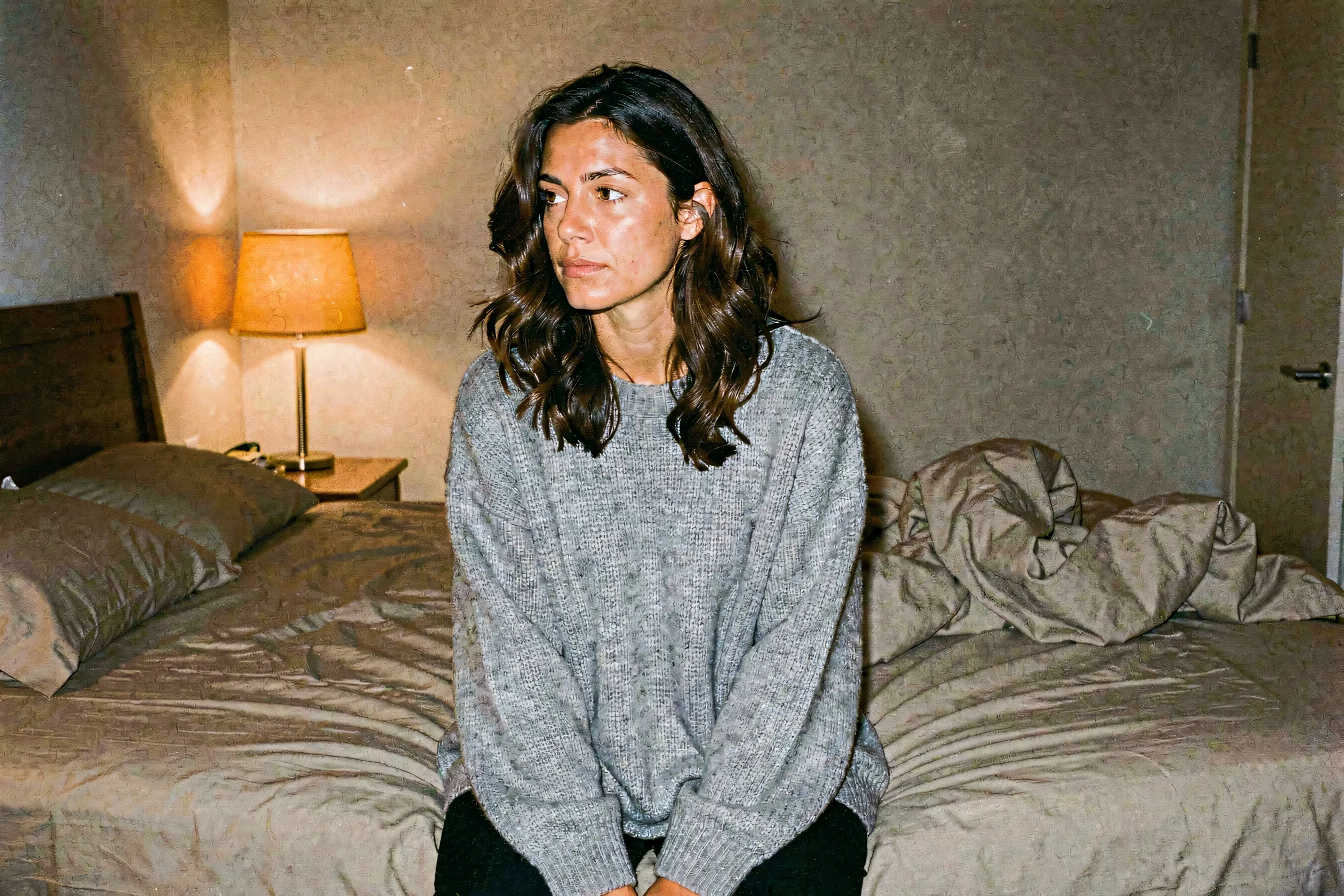 A woman in an oversized gray knit sweater sitting thoughtfully on the edge of an unmade bed in warm lamplight.