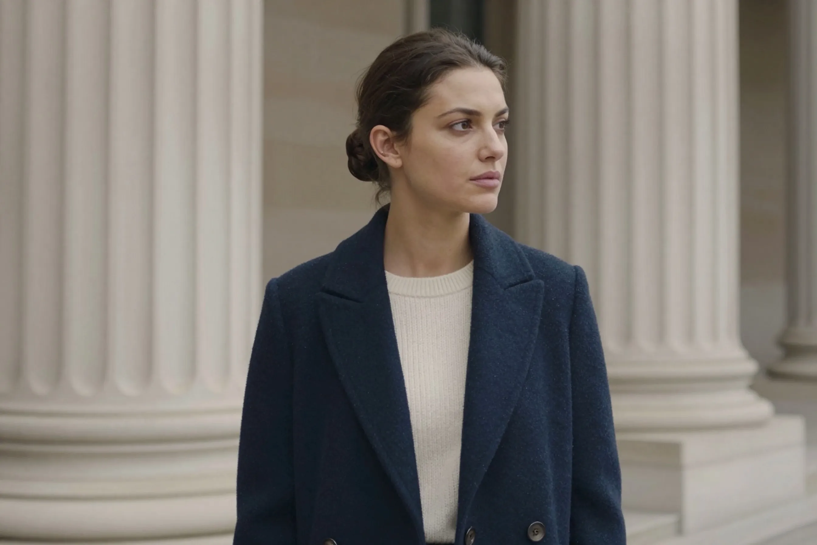 A woman in her early 30s with dark hair in a neat low bun wearing a tailored navy coat, standing beside classical stone pillars in an art museum courtyard.
