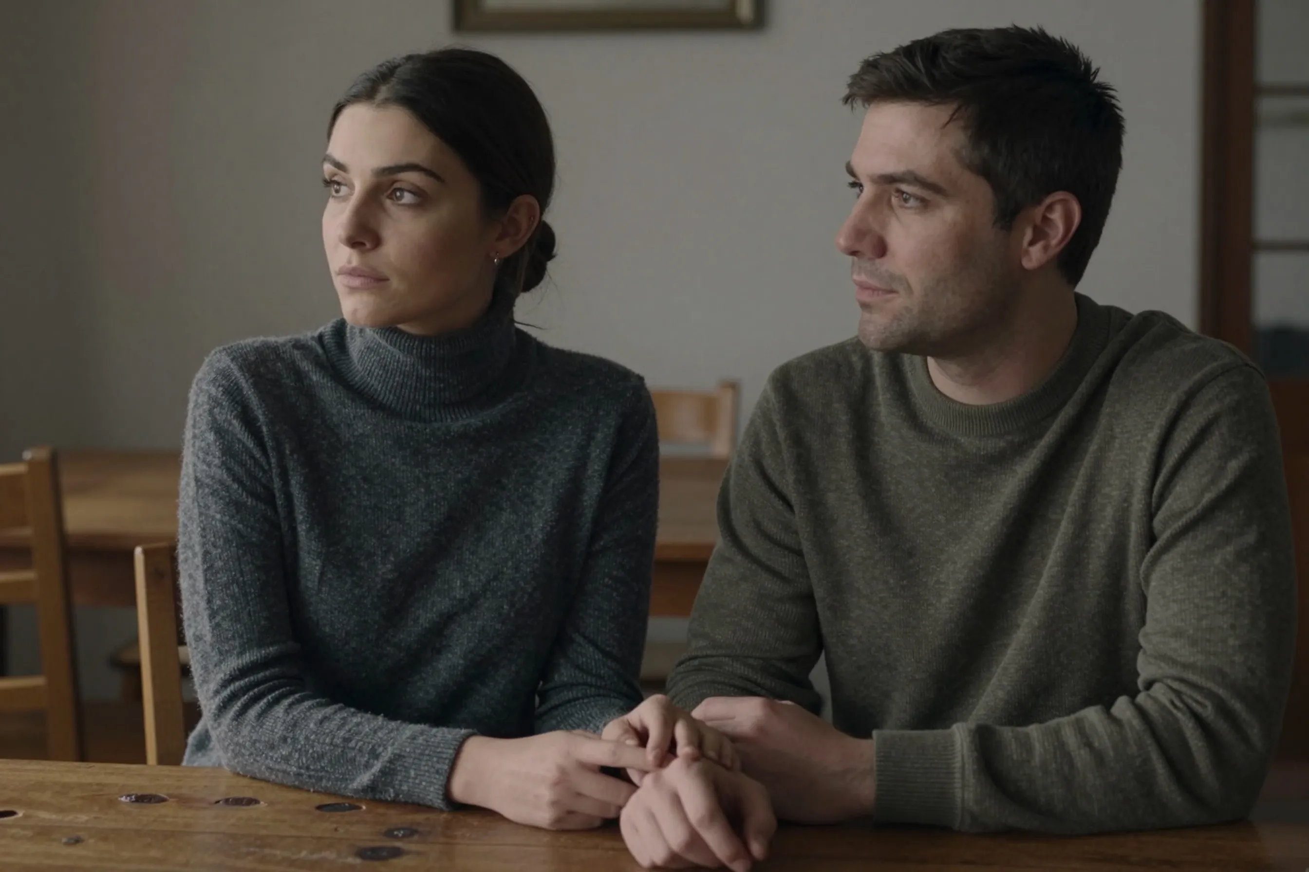 A woman in her early 30s and a man in his early 30s sitting at a wooden dining table at dusk with hands resting together in conversation.