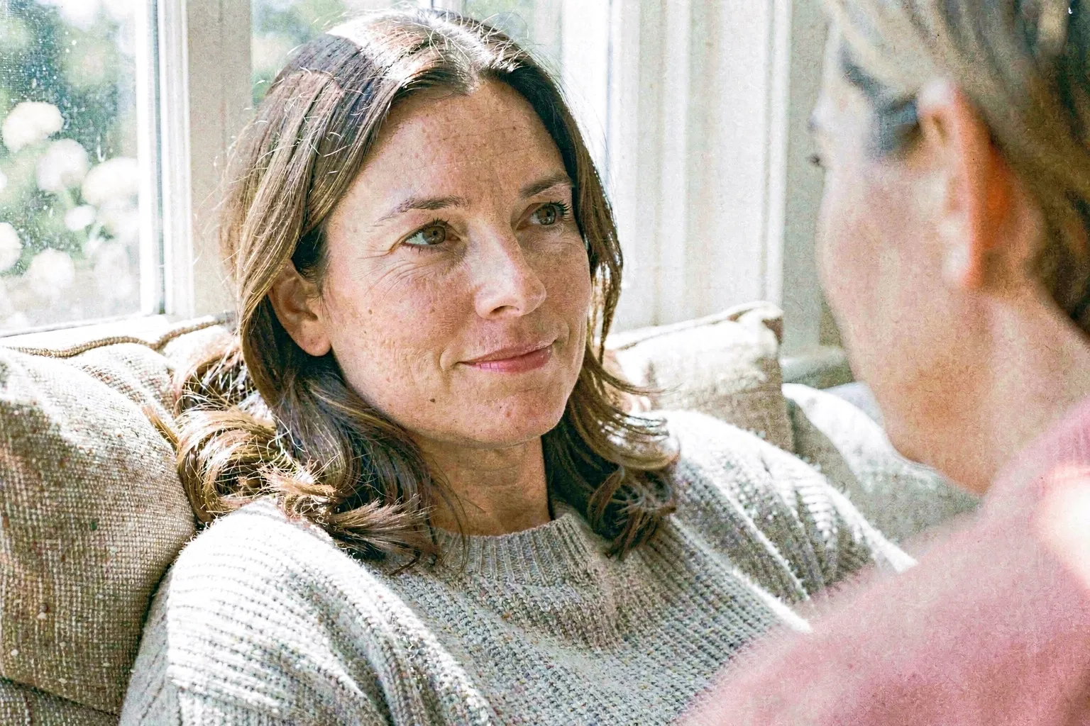 A close-up portrait of an adult woman resting comfortably against cushions by a bright window, her gaze thoughtful and alert with a subtle, affectionate half-smile