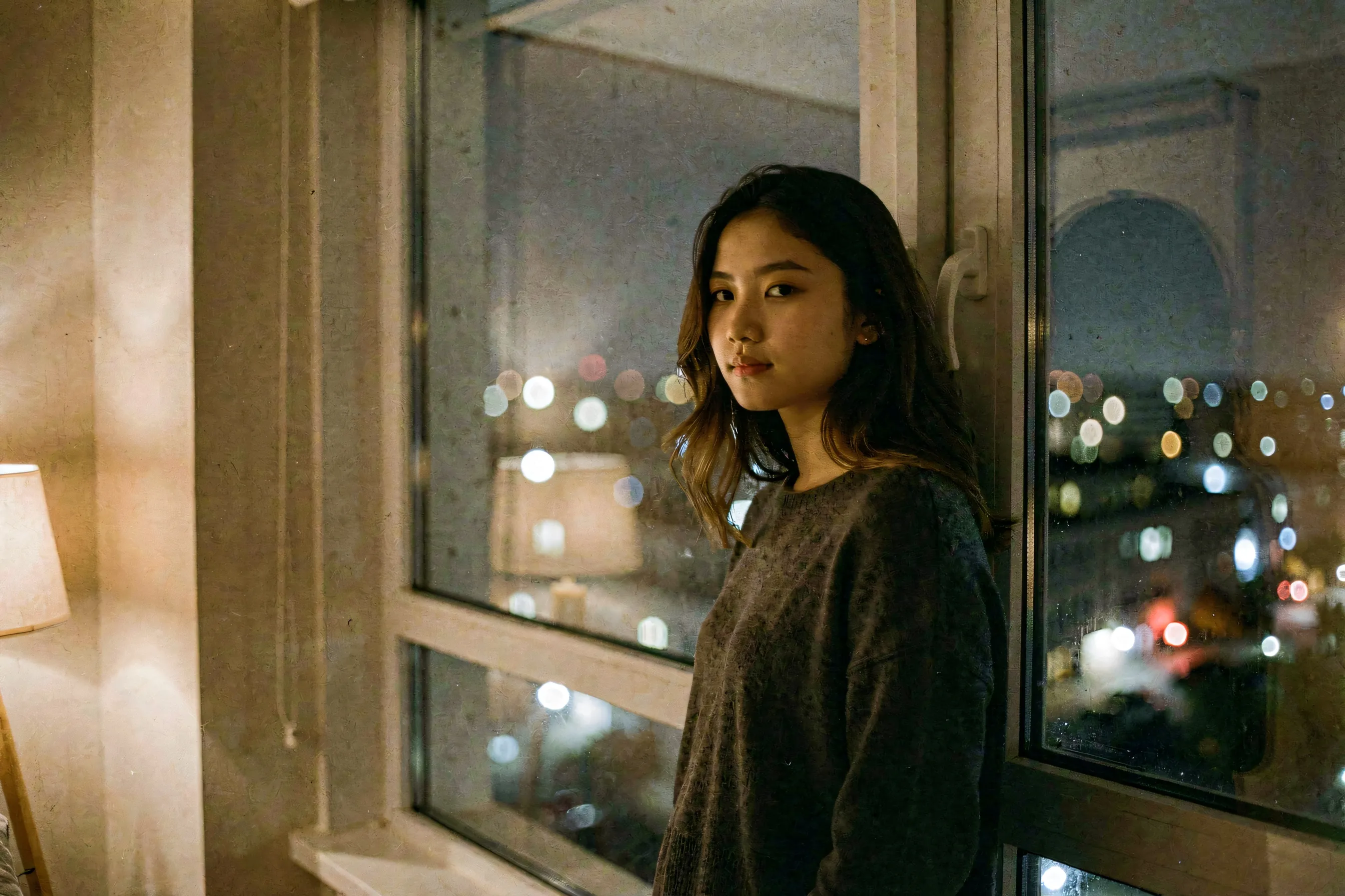 A young woman standing alone by a large apartment window at night, city lights blurring outside, her face calm and unreadable as she looks out