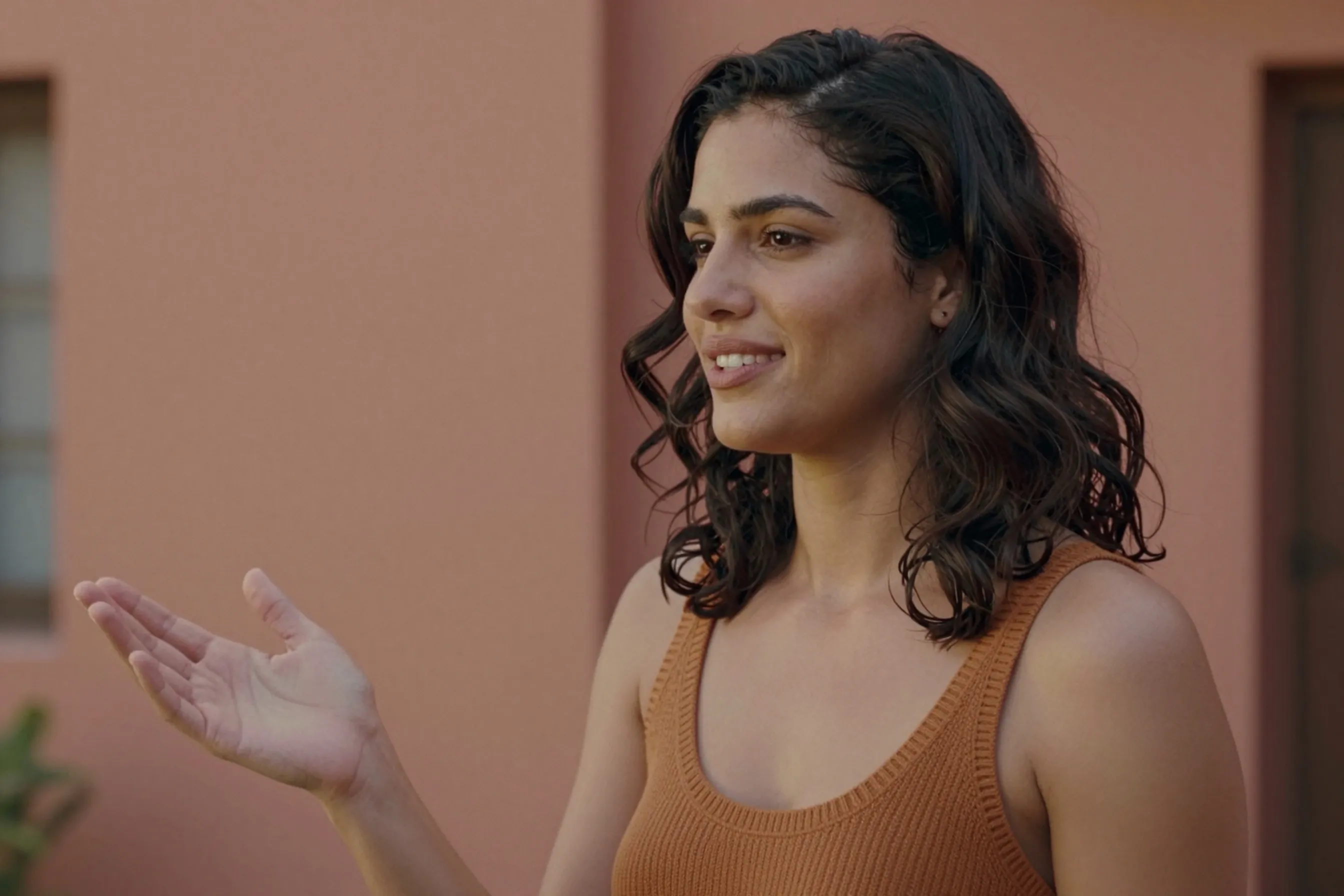 A woman in her late 20s with wavy dark hair wearing an ochre knit top, standing upright beside terracotta walls in afternoon light.