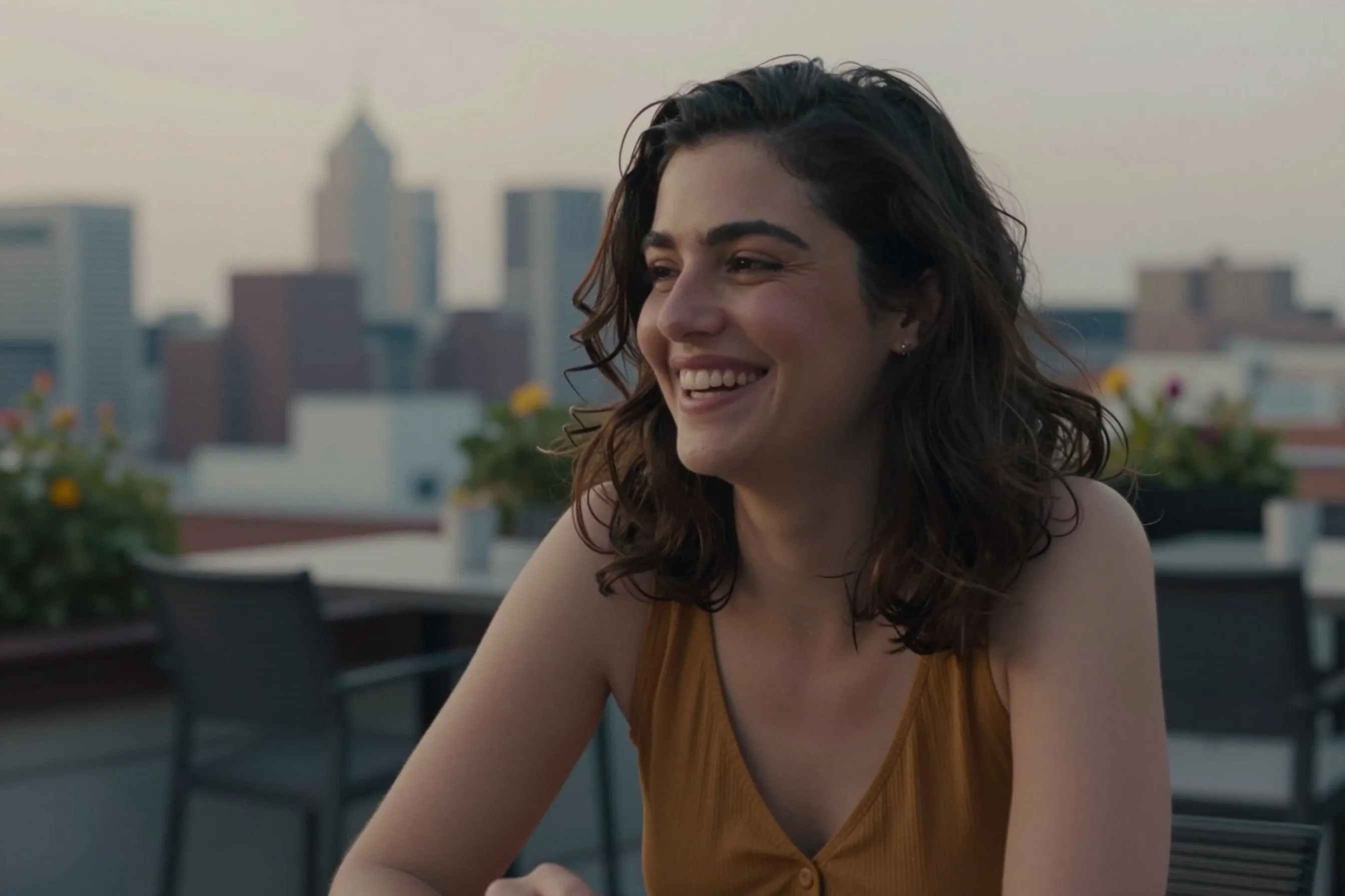 A woman in her early 30s with dark wavy hair wearing an ochre top, seated upright at an outdoor dining table overlooking the city skyline at sunset.