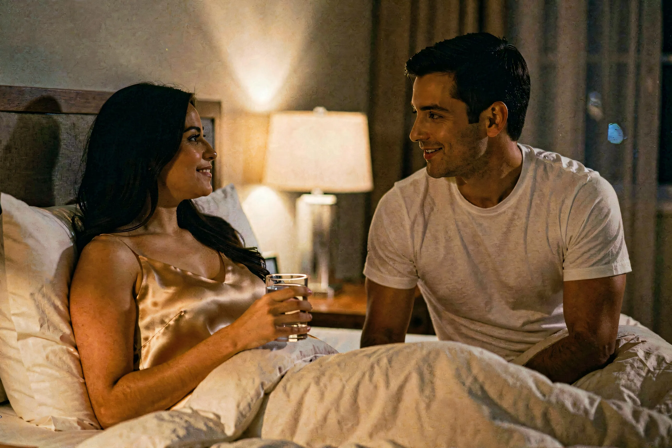 A woman propped against pillows in bed holding a glass of water, a man beside her talking and smiling