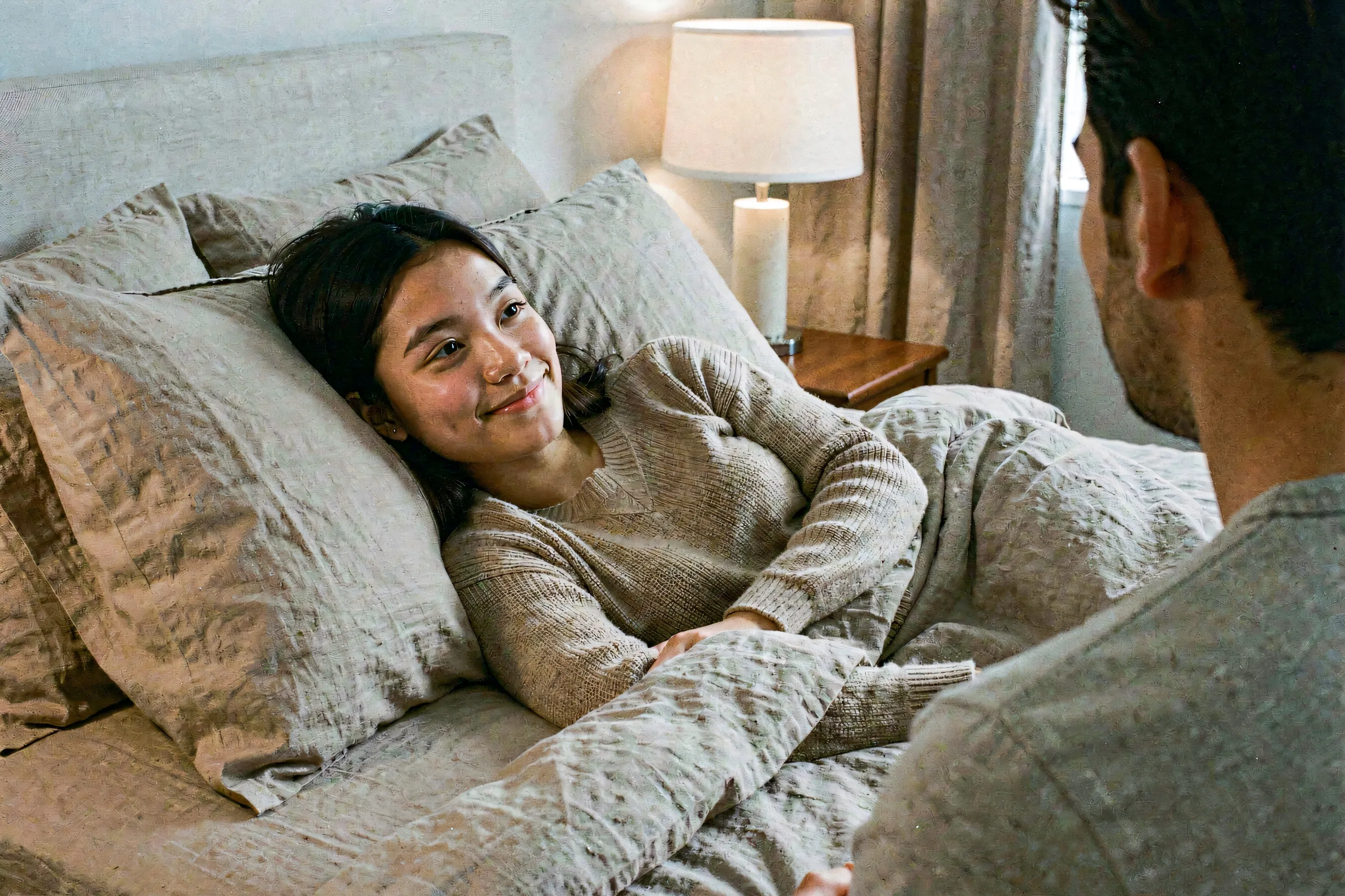 A woman reclining peacefully on a bed draped in soft neutral linens, warm lamplight catching her thoughtful expression as she gazes toward her partner