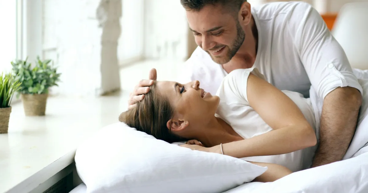 A couple playfully wrestling and laughing in bed, tangled in white sheets, morning light on their faces