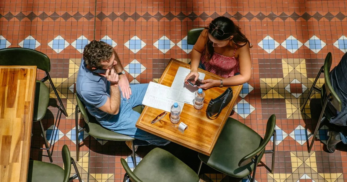 A man and woman sitting at a cafe table, him leaning back with his phone, his gaze drifting past her toward the door