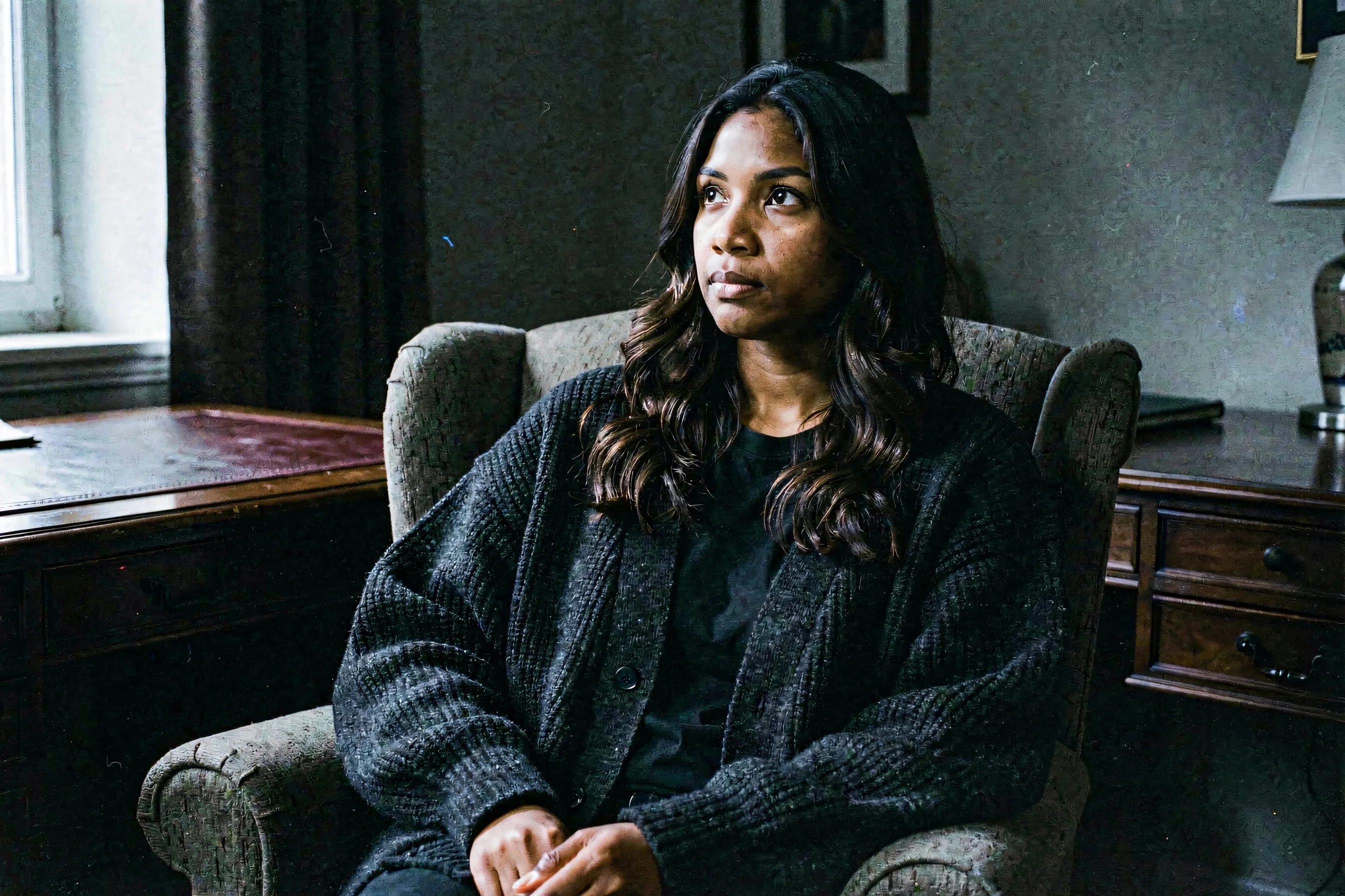 A woman with long dark wavy hair sitting in an armchair in moody low light, looking outward with quiet composure.