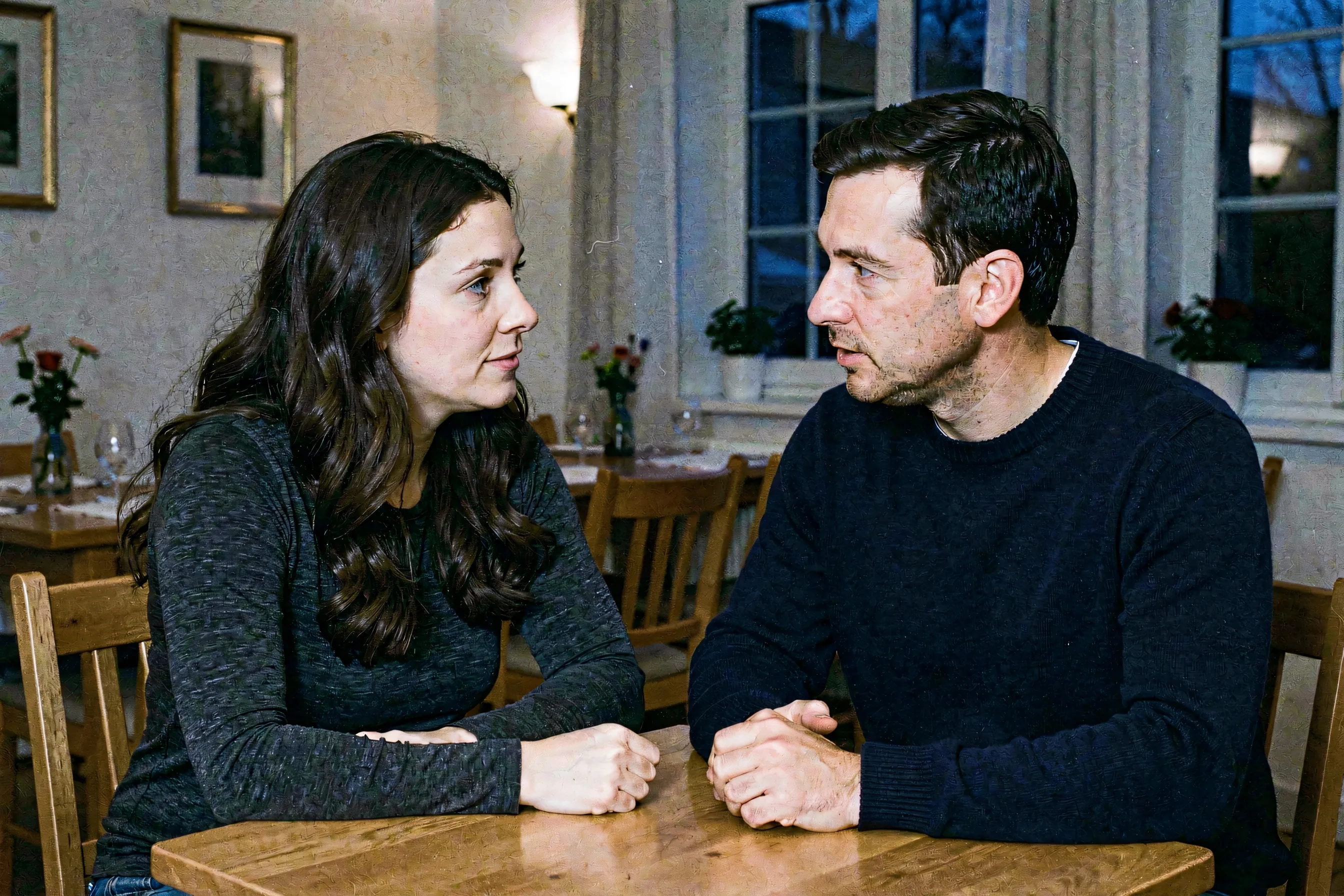 A woman with long dark wavy hair and her male partner sitting at a small table in dusk light, engaged in earnest conversation.
