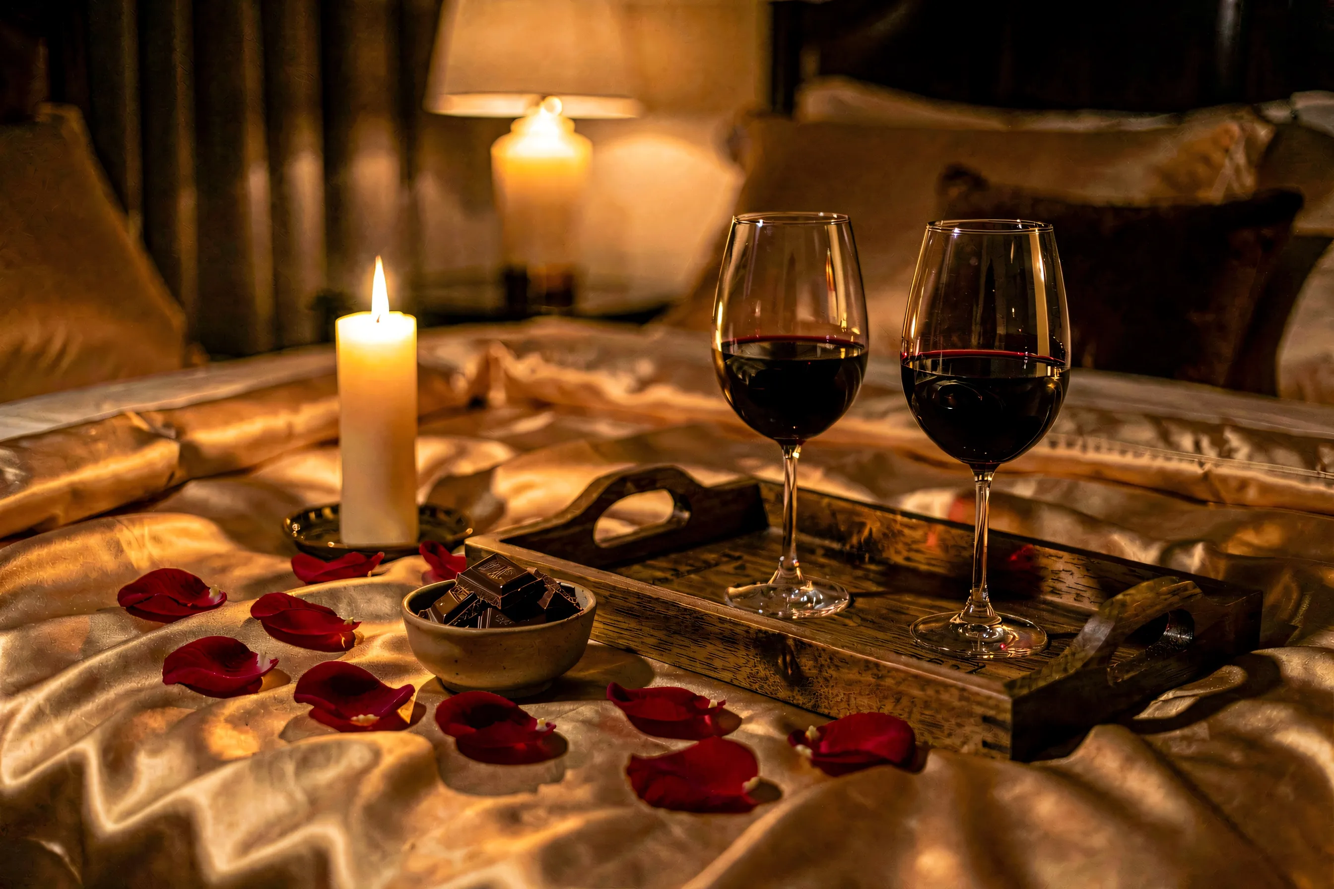 A candlelit bedroom at night with rose petals on the bed, a bowl of chocolate, two glasses of wine, and silk sheets